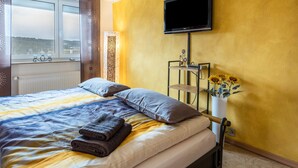 A hotel room with a bed, a TV mounted on the wall, a window with a view, and a vase with flowers on a side table. - Apartment 'Schwarzwaldblick' with Mountain View, Balcony, and Wi-Fi (St.Georgen im Schwarzwald)