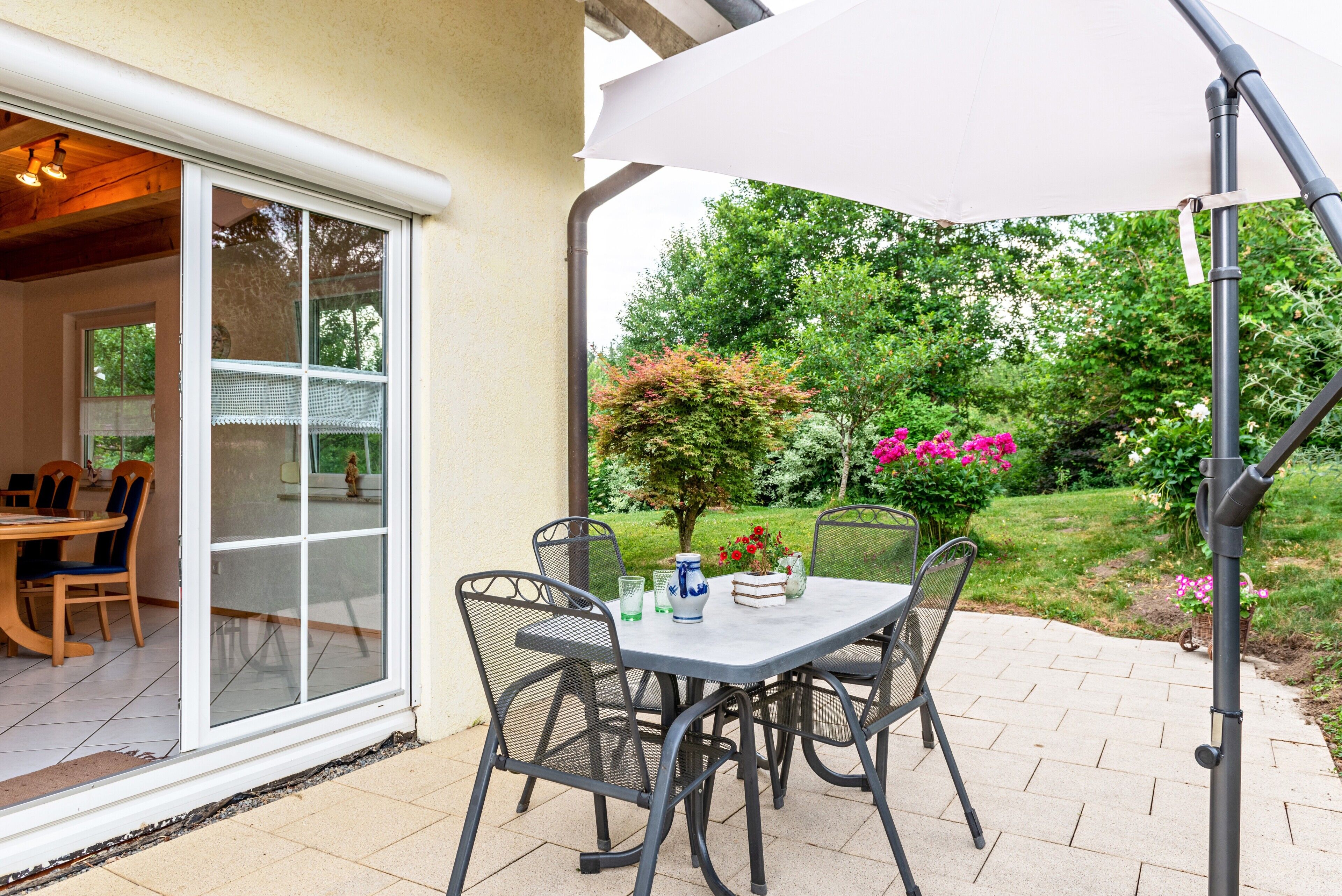 Eine Terrasse im Freien mit Tisch und Stühlen, einem weißen Sonnenschirm und Blick auf einen Garten.