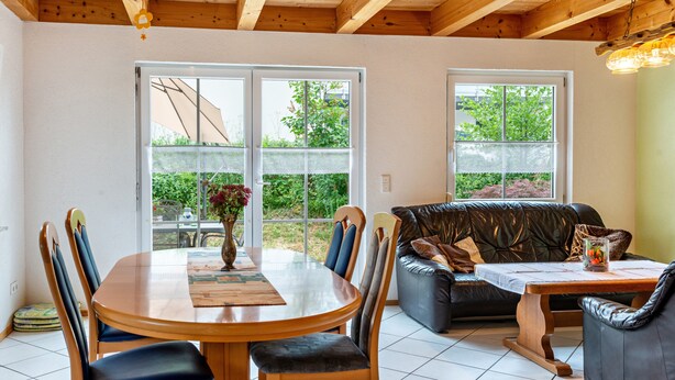Ein Essbereich mit Holztisch und Stühlen, einer schwarzen Ledersofa und großen Fenstern mit Blick auf Grünflächen.