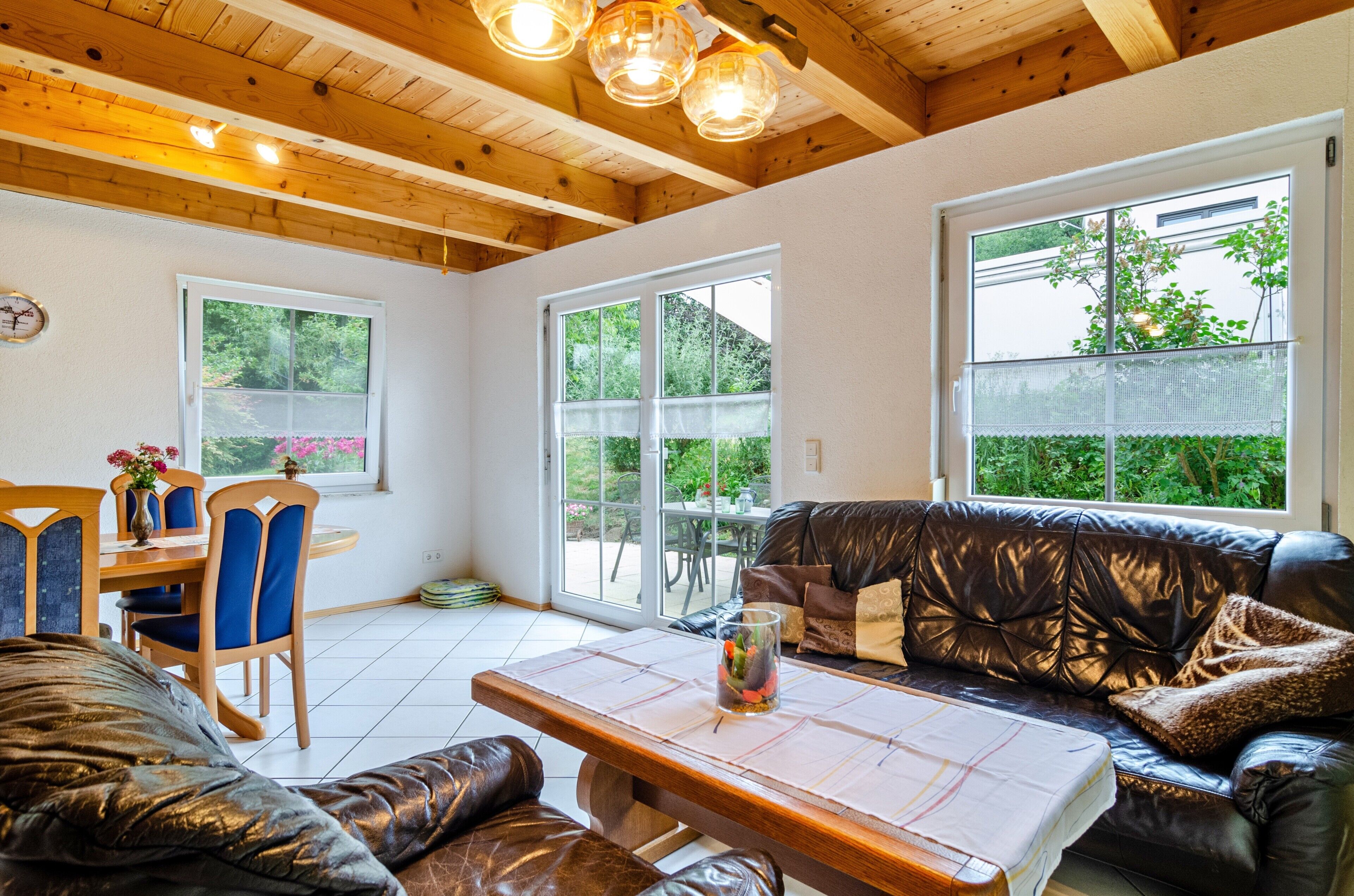 A cozy interior with a wooden ceiling, a dining area with blue chairs, a leather sofa, and large windows offering a view of greenery.