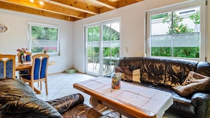 Ein gemütliches Interieur mit Holzbalkendecke, einer Essecke mit blauen Stühlen, einem Ledersofa und großen Fenstern mit Blick auf viel Grün.