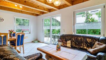 A cozy interior with a wooden ceiling, a dining area with blue chairs, a leather sofa, and large windows offering a view of greenery.
