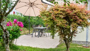 Eine Terrasse im Freien mit Tisch und Stühlen, umgeben von viel Grün und einem Baum.