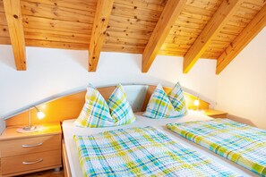 Une chambre avec un plafond en bois, un lit recouvert d’une couette à carreaux, une table de chevet avec une lampe et une commode.