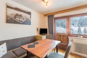 Une pièce chaleureuse avec une table en bois, un canapé, un téléviseur et une fenêtre donnant sur un paysage enneigé.