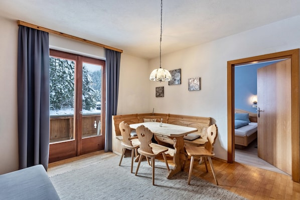 A dining area with a wooden table and chairs, a hanging light fixture, and a view of snow-covered trees through a window.