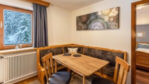 Une pièce d’angle avec une table en bois, des sièges rembourrés, un radiateur, une fenêtre donnant sur des arbres couverts de neige et un tableau abstrait accroché au mur.
