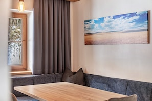 Ein Zimmer mit Holztisch, grauem Sofa, einem Fenster mit Vorhängen und einem Landschaftsbild an der Wand.