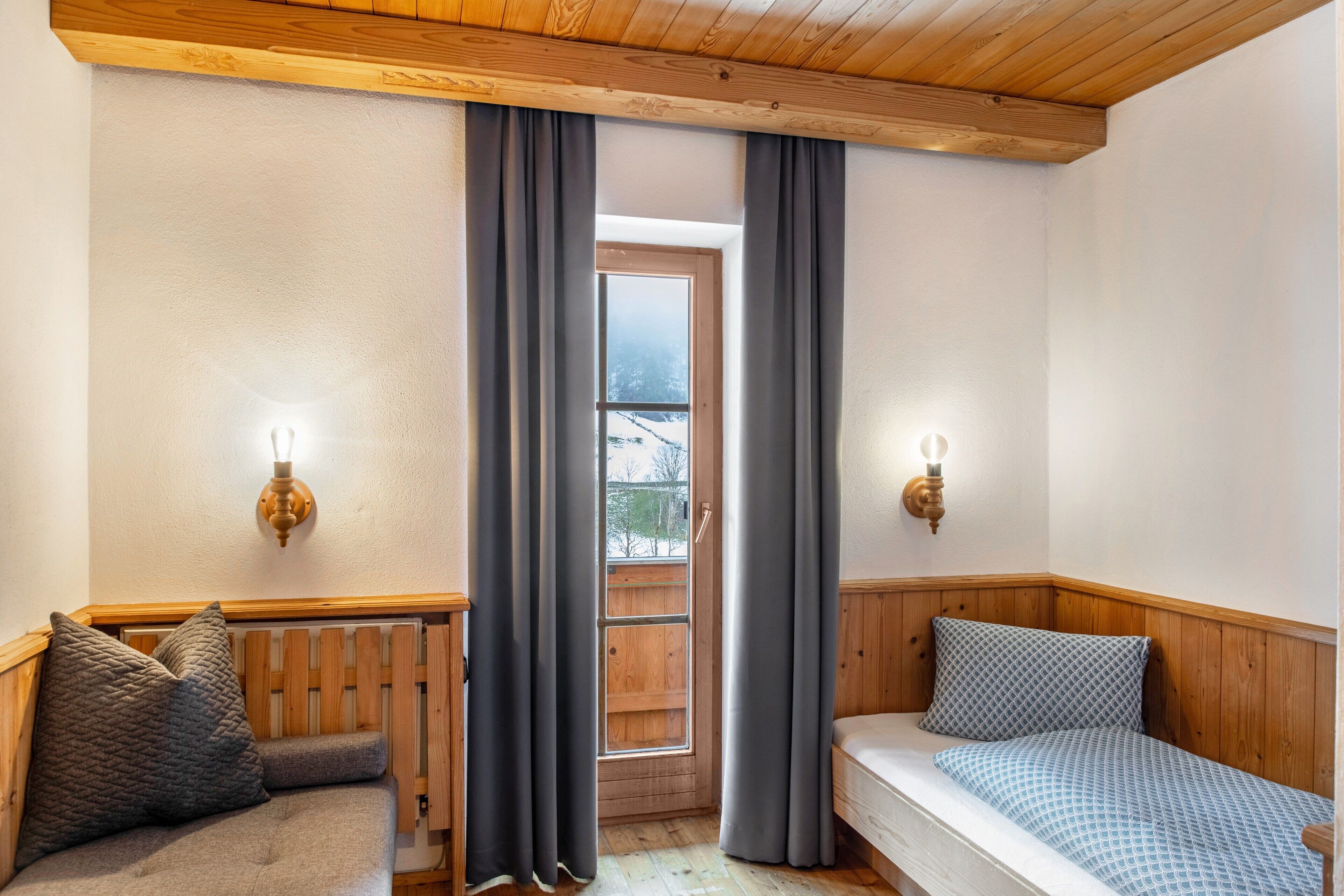 Une pièce chaleureuse avec un plafond en bois, une fenêtre avec des rideaux, un radiateur et un lit recouvert d’une literie à motifs.