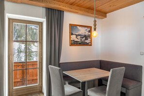 A dining area with a wooden table and grey upholstered booth seating, a framed picture on the wall, and a window with a view of trees.