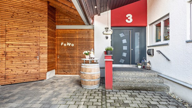 Une façade en bois avec une porte rouge portant le numéro « 3 » et un petit tonneau décoré de fleurs.