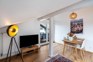 A modern living room with a flat-screen TV, a tripod lamp, a dining table with chairs, and a large window offering a view of the outside.