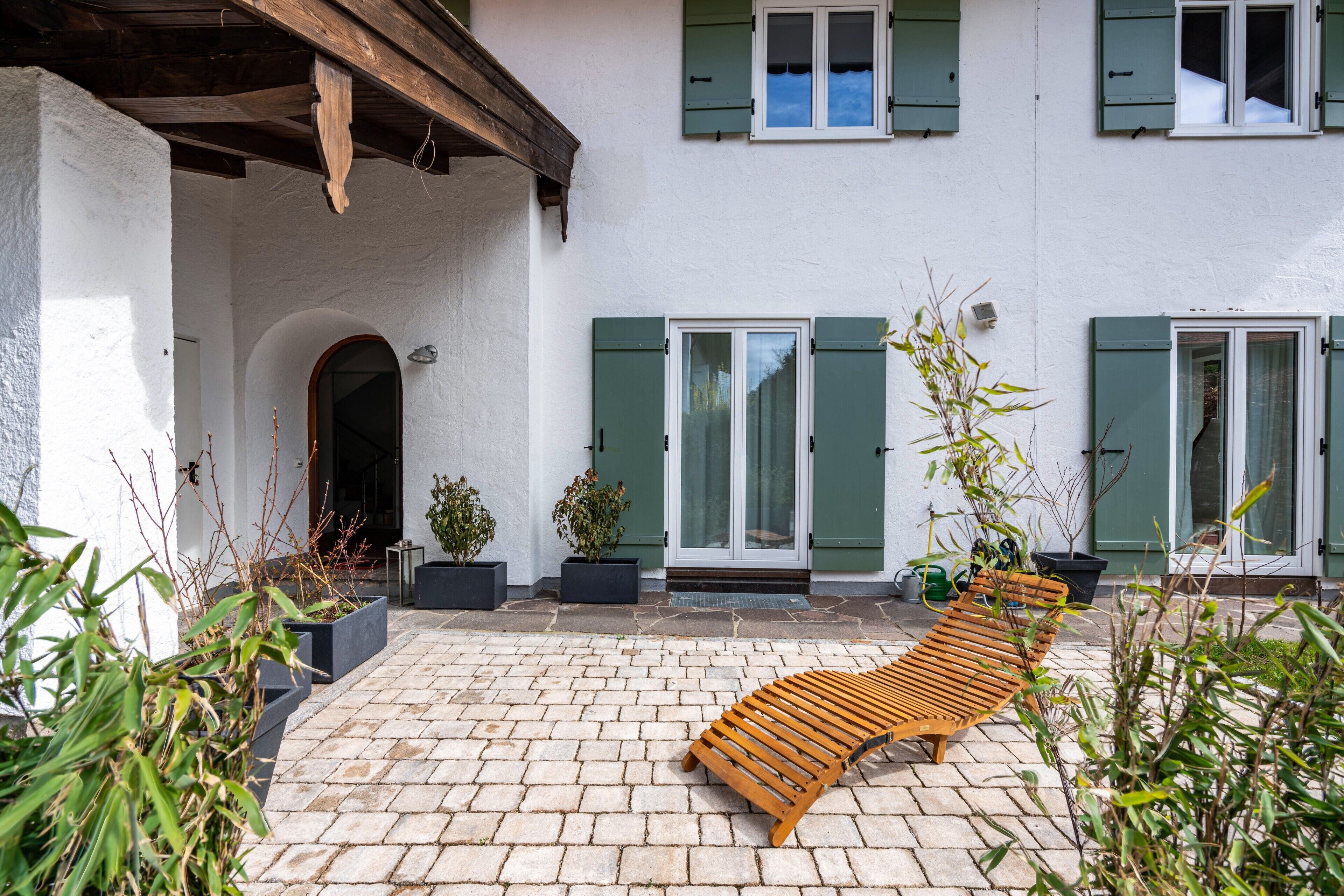 Ein Innenhof mit einer Holzbank, Blumentöpfen und einem Gebäude mit grünen Fensterläden und weißen Wänden.