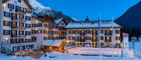 A multi-story hotel with balconies, a wooden cabin, and a snowy landscape with mountains in the background.