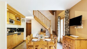 Una mesa de comedor de madera con sillas, una cocina con armarios de madera y un microondas, una escalera y un televisor de pantalla plana montado en la pared.