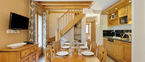 Ein Essbereich mit einem Holztisch, gedeckt für sechs Personen, der von Holzkästen und einem Fernseher links eingerahmt wird. Eine Treppe führt zu einer oberen Ebene.