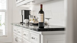 A kitchen counter with a wine bottle and glass, white cabinets, and a coffee machine. - Orange Blossom Cottage Near Sequoias w/EV Charger (Exeter)