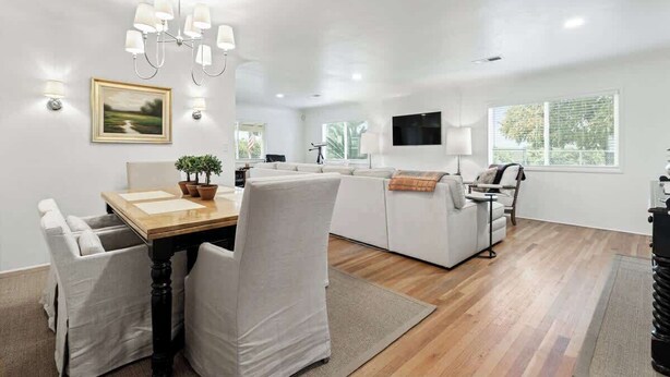 A modern living room with a wooden dining table, white sofas, a TV mounted on the wall, and a chandelier. - Orange Blossom Cottage Near Sequoias w/EV Charger (Exeter)