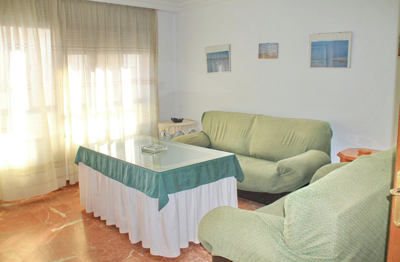 A living room with a glass-top table, a green sofa, a patterned armchair, and a small table with a bowl.