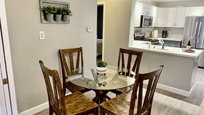 Un comedor con una mesa de vidrio rodeada de sillas de madera, una pequeña planta en maceta en la pared y una cocina al fondo con armarios blancos y electrodomésticos de acero inoxidable.