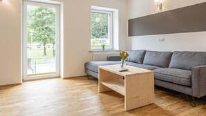 A modern living room with a grey sofa, a wooden coffee table, and two windows providing a view of greenery outside.