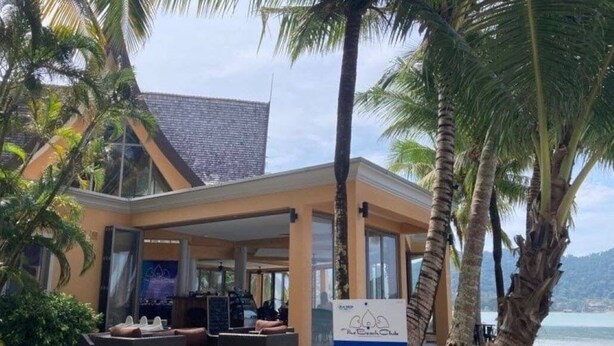 Um hotel na praia com área de estar coberta ao ar livre, palmeiras e uma placa indicando ser um restaurante de dia inteiro com cozinha ocidental e tailandesa.
