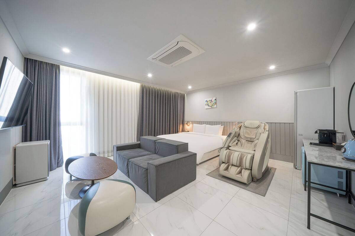 Una habitación de hotel moderna con cama, sillón de masaje, zona de estar y una pequeña mesa.