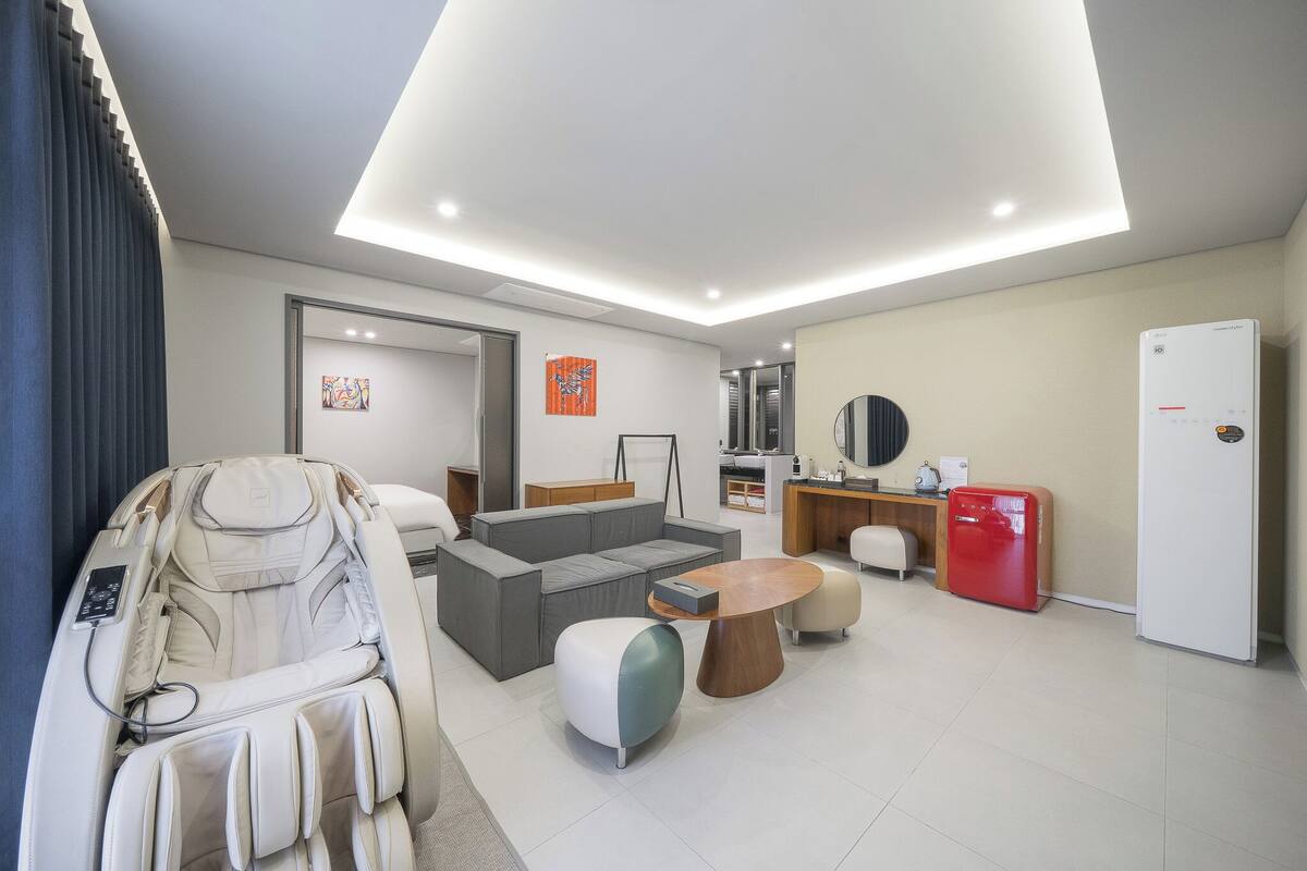 Una sala moderna con sillón de masaje, sofá gris, mesa de centro de madera y un refrigerador rojo.