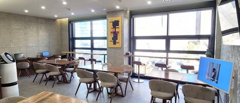 A modern cafe with tables and chairs, large windows, and a wall-mounted screen displaying a video.