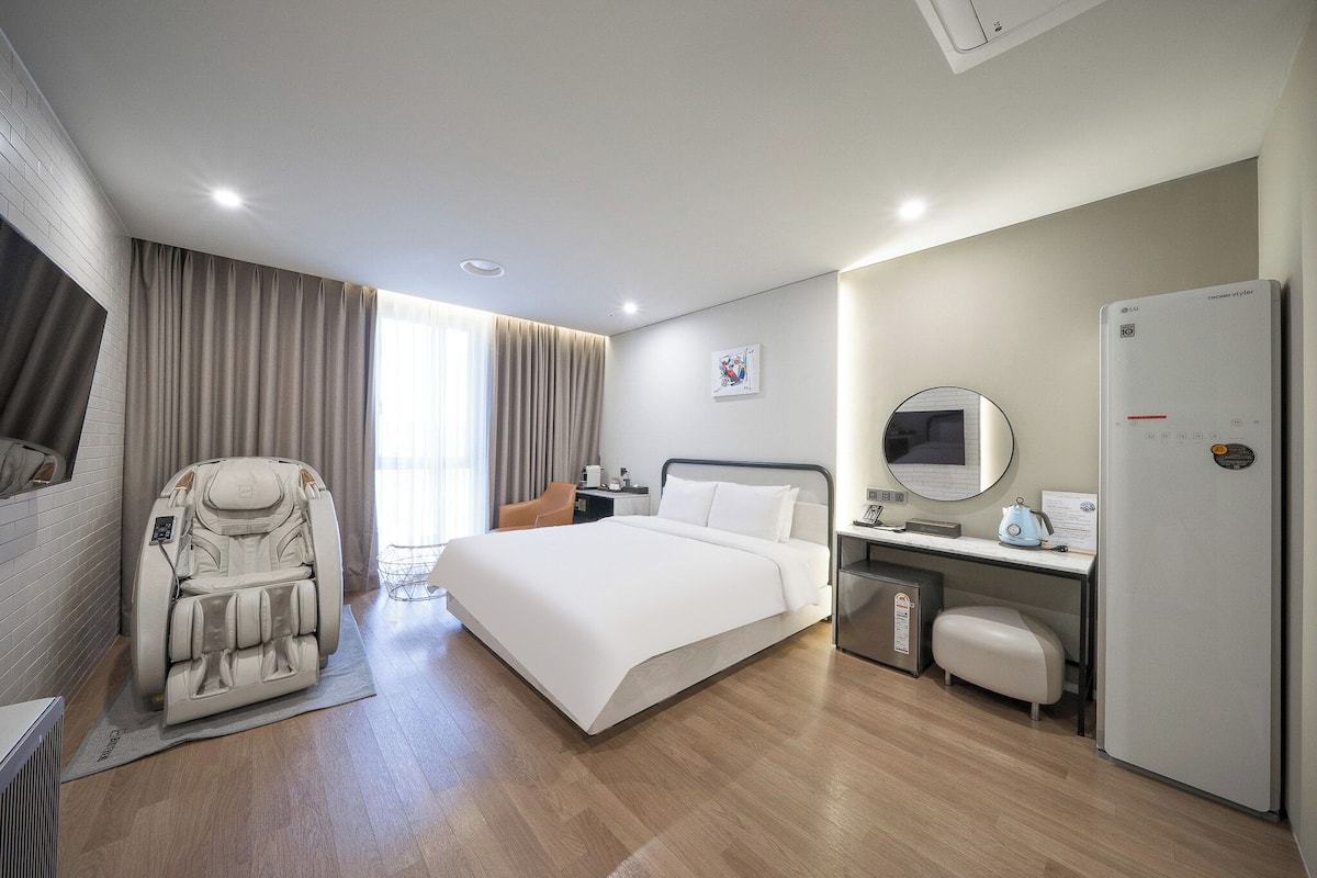 Habitación de hotel con cama, sillón de masajes, escritorio con computadora, refrigerador y ventana con cortinas.