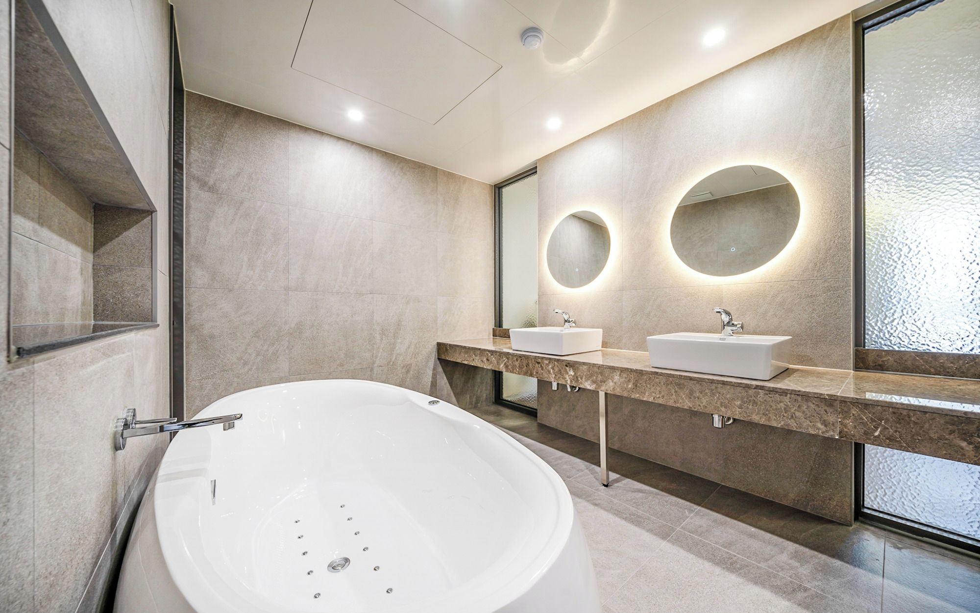 A modern bathroom with a freestanding bathtub, two sinks, and a large mirror with circular lighting.