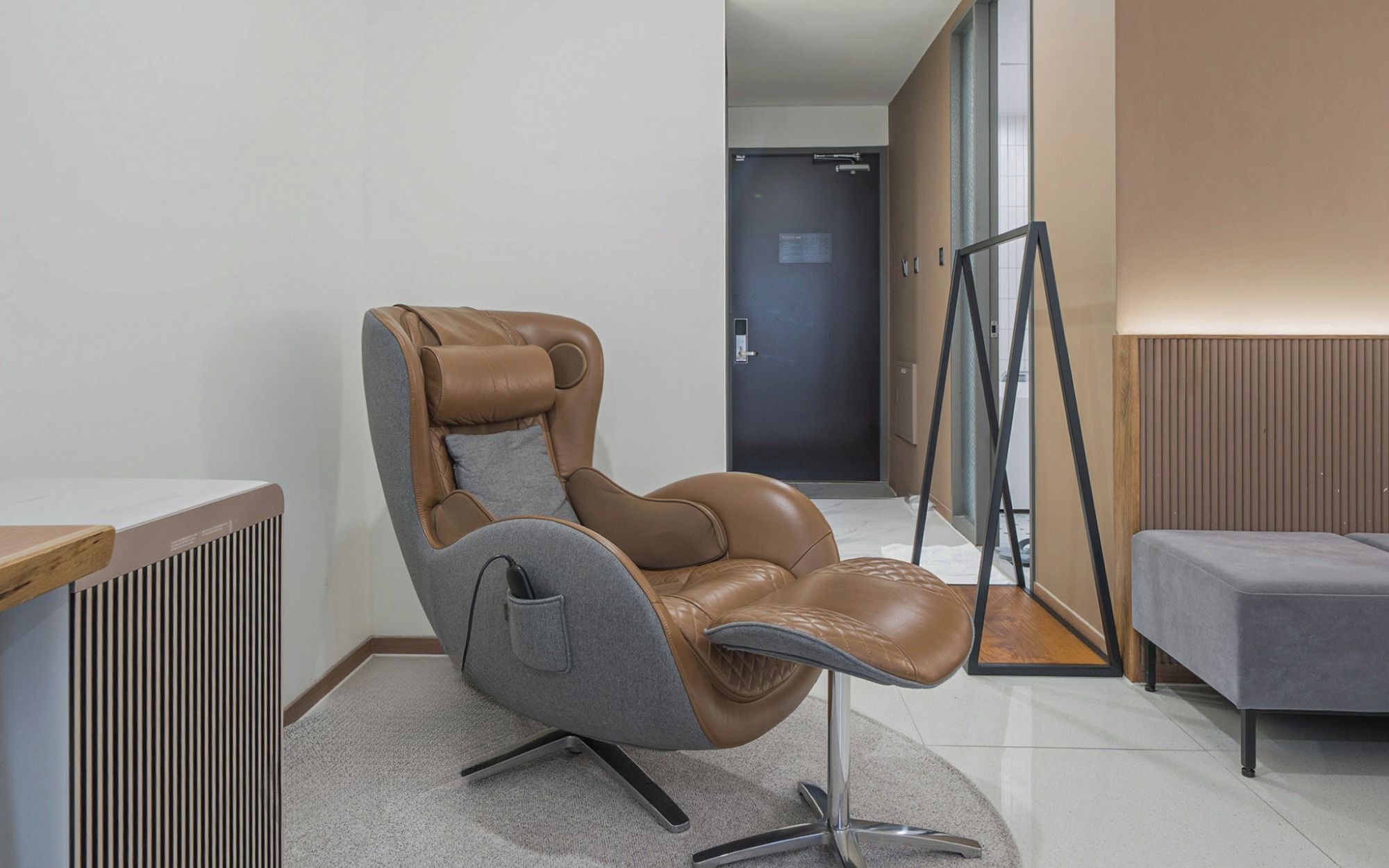 Una moderna silla de masaje en una habitación de hotel con suelo de madera, un sofá gris y una puerta al fondo.