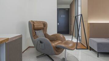 Una moderna silla de masaje en una habitación de hotel con suelo de madera, un sofá gris y una puerta al fondo.