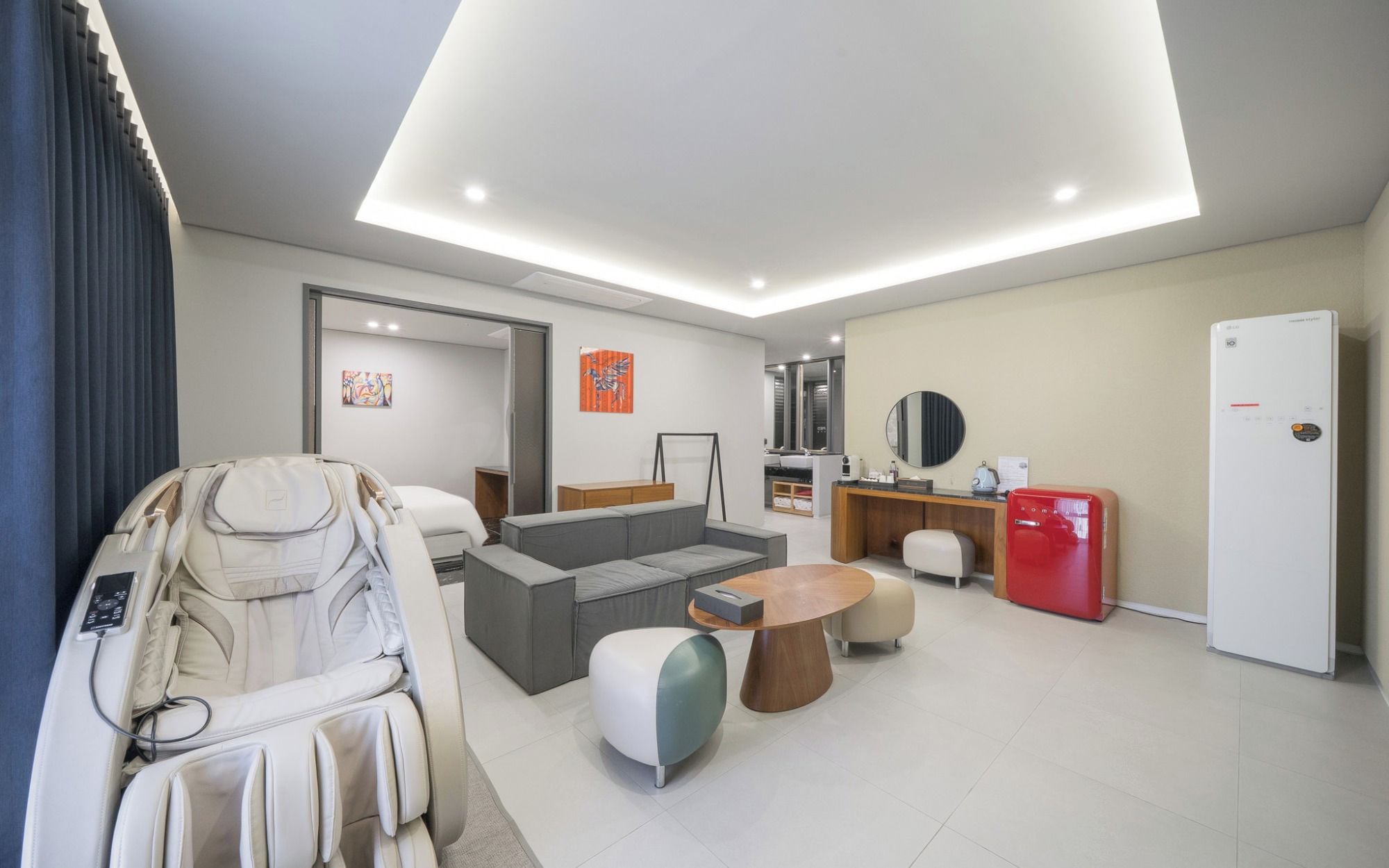 Et moderne hotelværelse med en massagestol, en sofa, et lille spisebord, et kompakt tekøkken og et stort spejl.