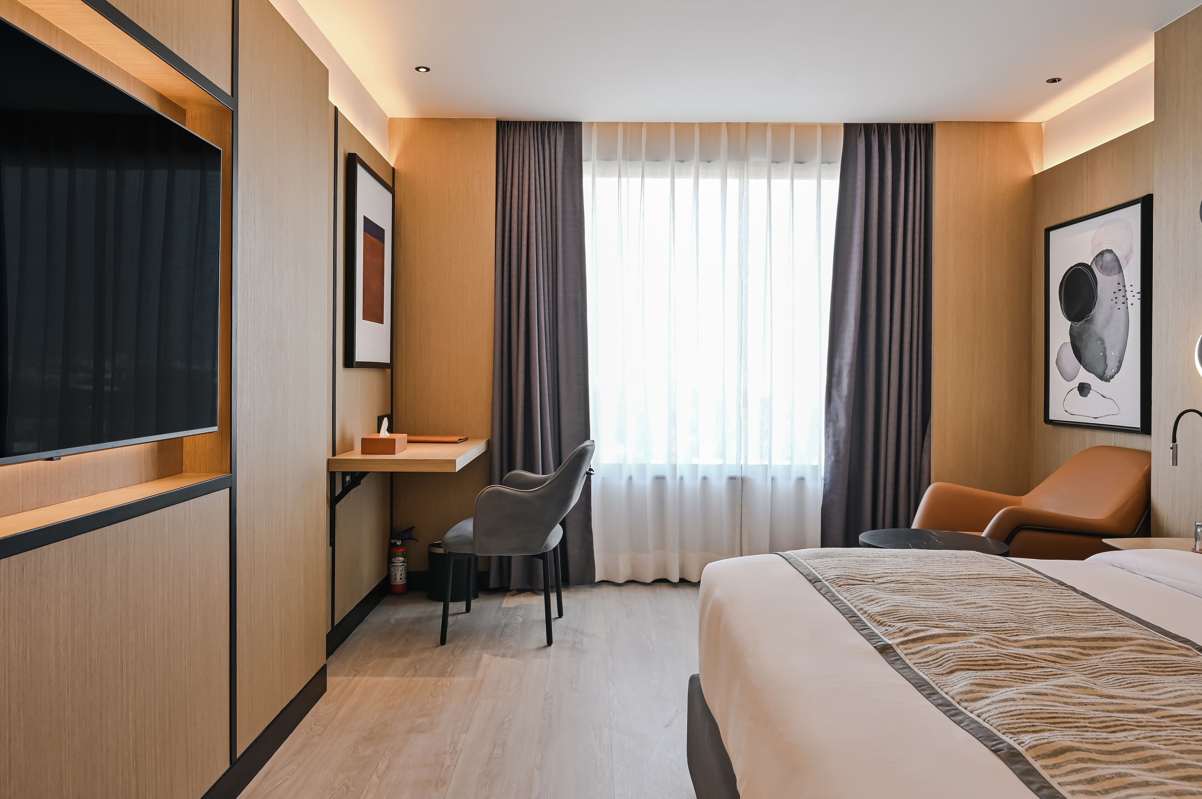 A hotel room with a bed, a desk with a chair, a TV, and a window with curtains.