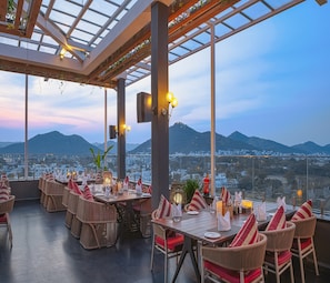 A restaurant with wicker chairs and tables, set against a backdrop of mountains and a cityscape.