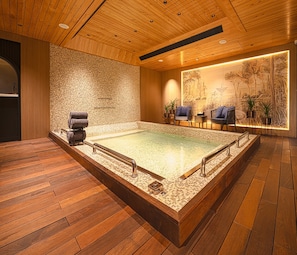 A spa room with a large hot tub, wooden flooring, a mosaic wall, and a seating area with plants.