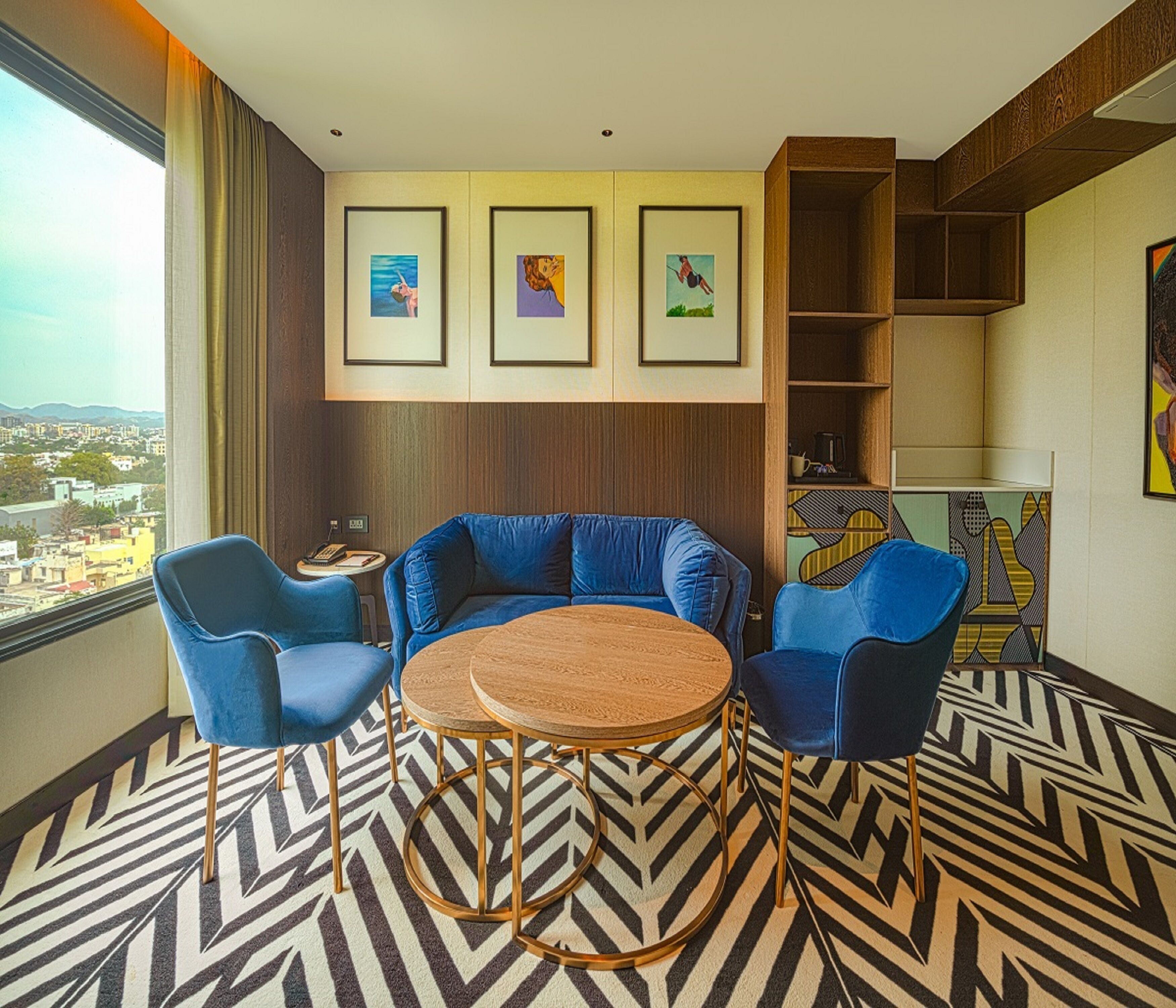 Een moderne hotelkamer met een groot raam, een blauwe zithoek, een ronde houten tafel en een vloer met een patroon.