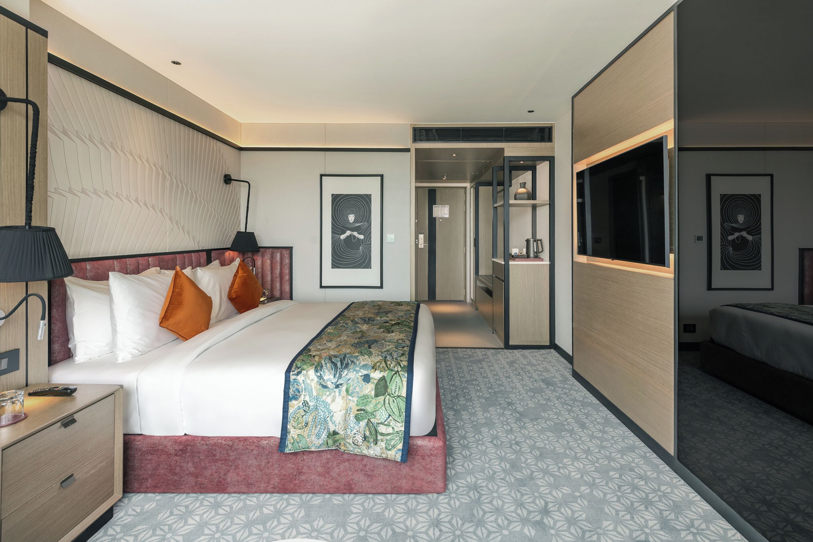 A modern hotel room with a large bed, a desk with a lamp, a wardrobe, and a bathroom visible through an open door.
