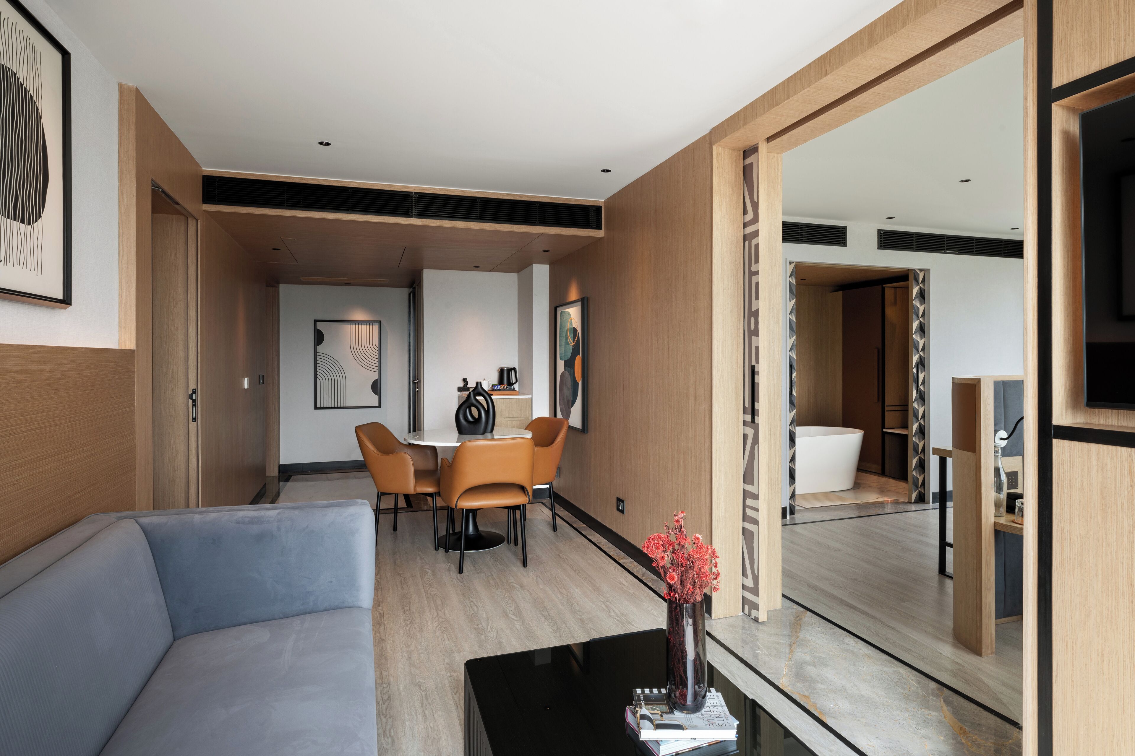Quarto de hotel moderno com sofá, área de refeições com mesa e cadeiras, banheiro com banheira e um espelho grande.