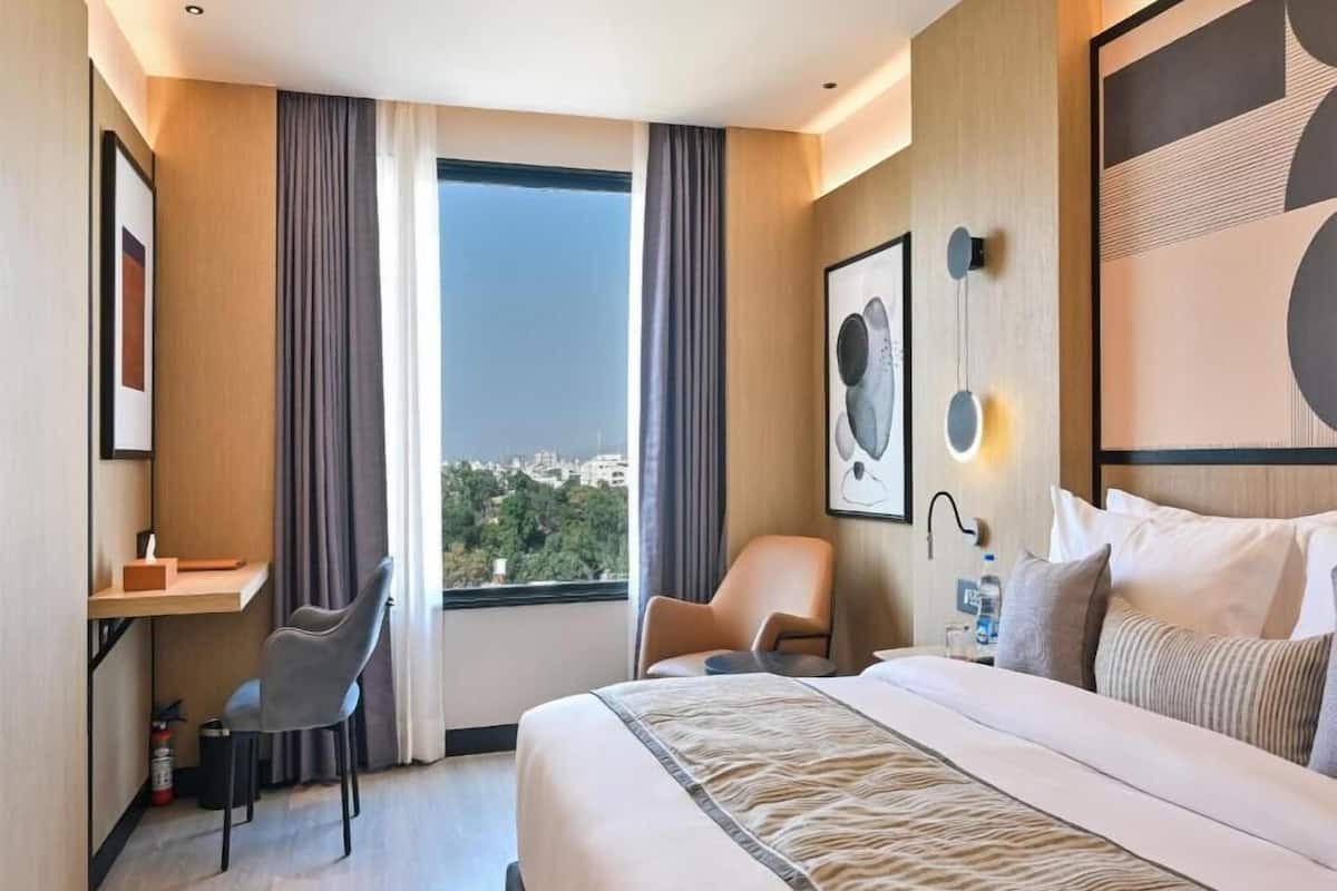 A hotel room with a bed, a desk, a chair, a large window with curtains, and artwork on the walls.