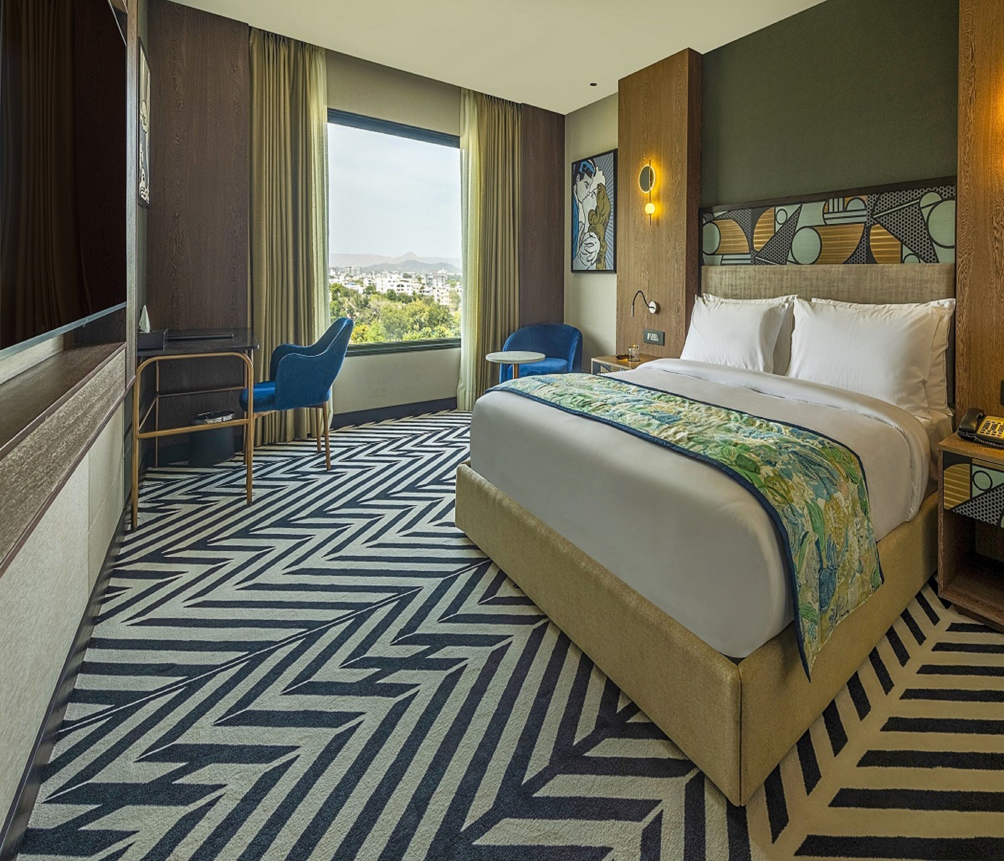 A hotel room with a large bed, a desk, a chair, and a view of the city.