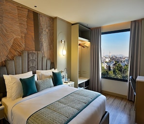 A hotel room with a large bed, a nightstand, a lamp, a view of the city through a window, and a wardrobe.
