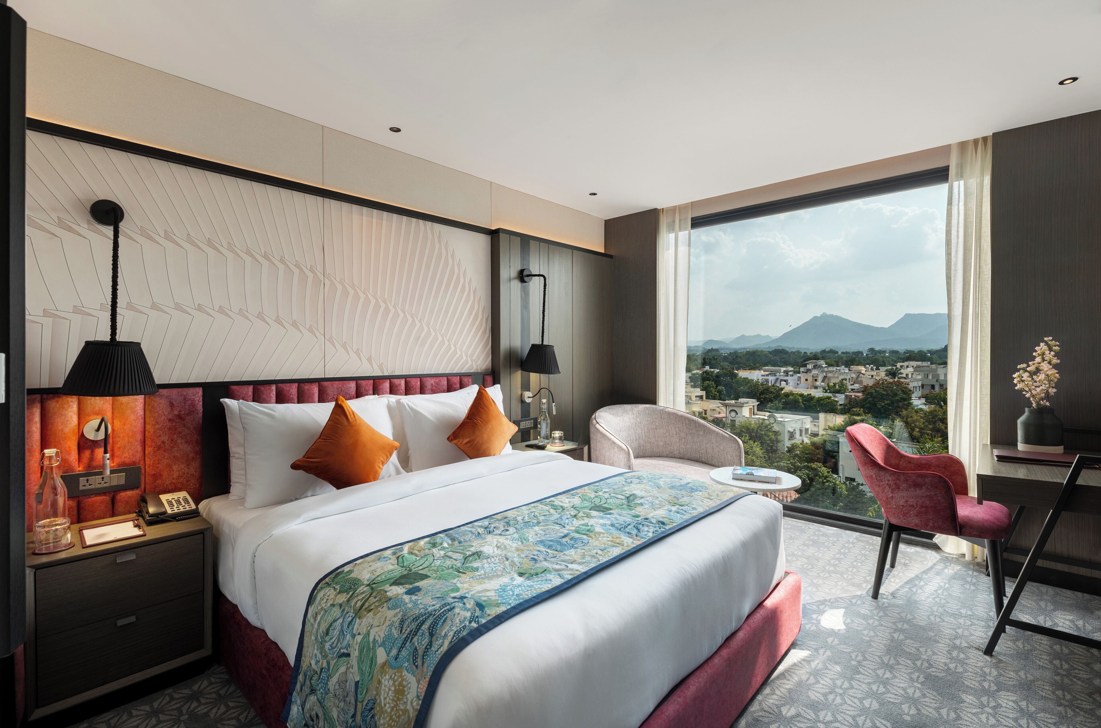 Habitación de hotel con una cama grande, un escritorio, una silla y vistas a un paisaje urbano a través de un ventanal amplio.
