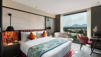 Habitación de hotel con una cama grande, un escritorio, una silla y vistas a un paisaje urbano a través de un ventanal amplio.