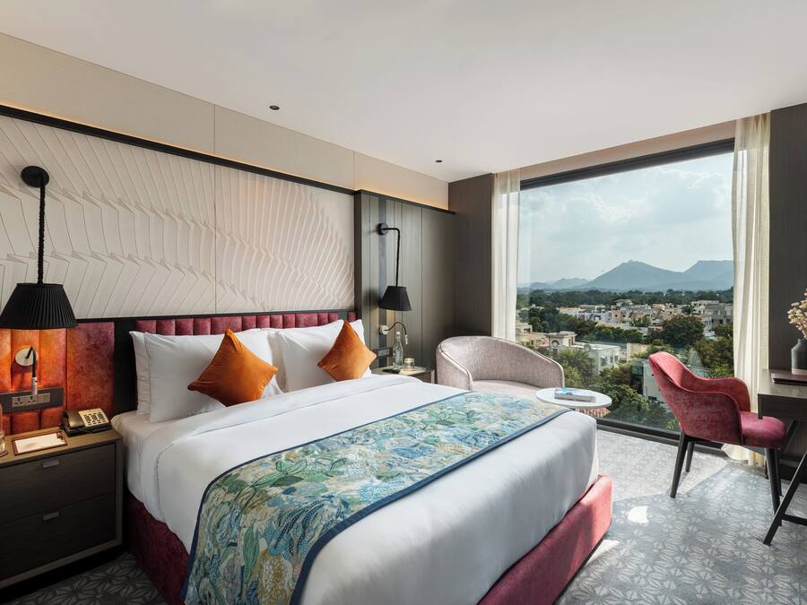 Habitación de hotel con una cama grande, un escritorio, una silla y vistas a un paisaje urbano a través de un ventanal amplio.