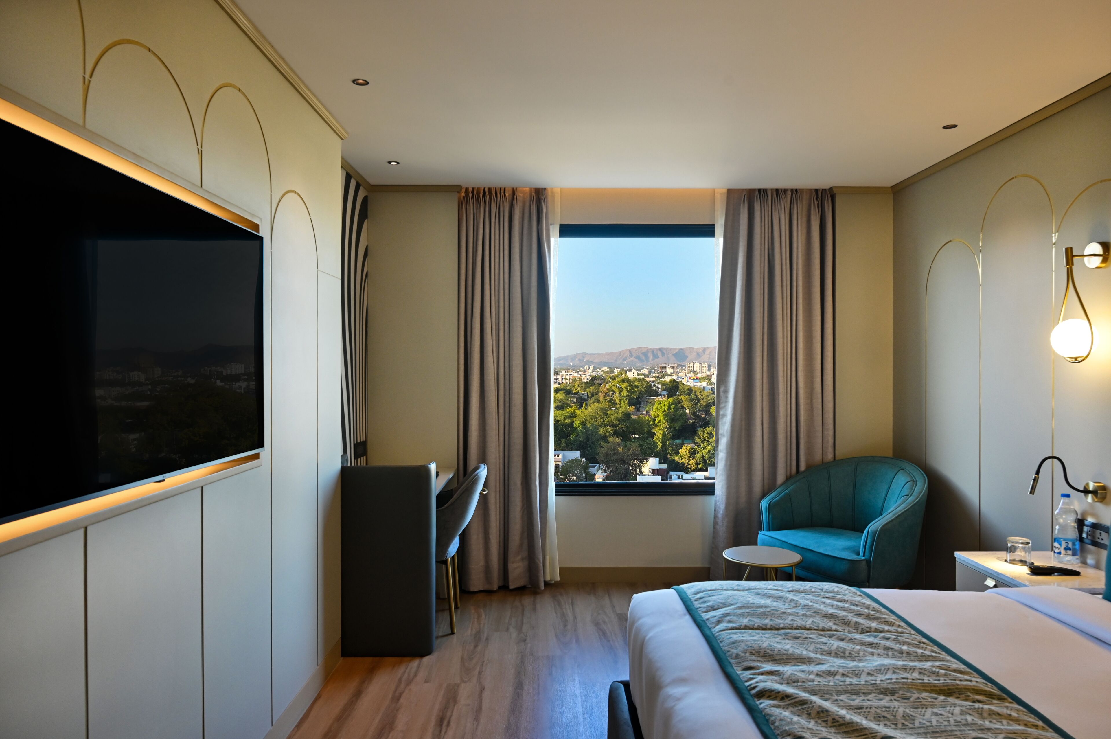 A hotel room with a bed, a desk, a chair, a TV, a window with curtains, and a view of a cityscape.