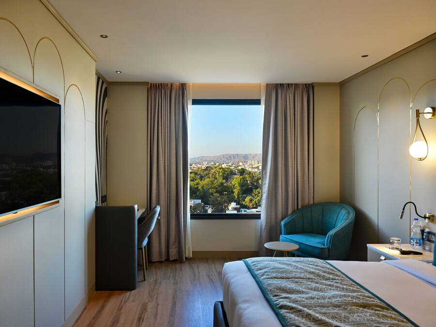 A hotel room with a bed, a desk, a chair, a TV, a window with curtains, and a view of a cityscape.