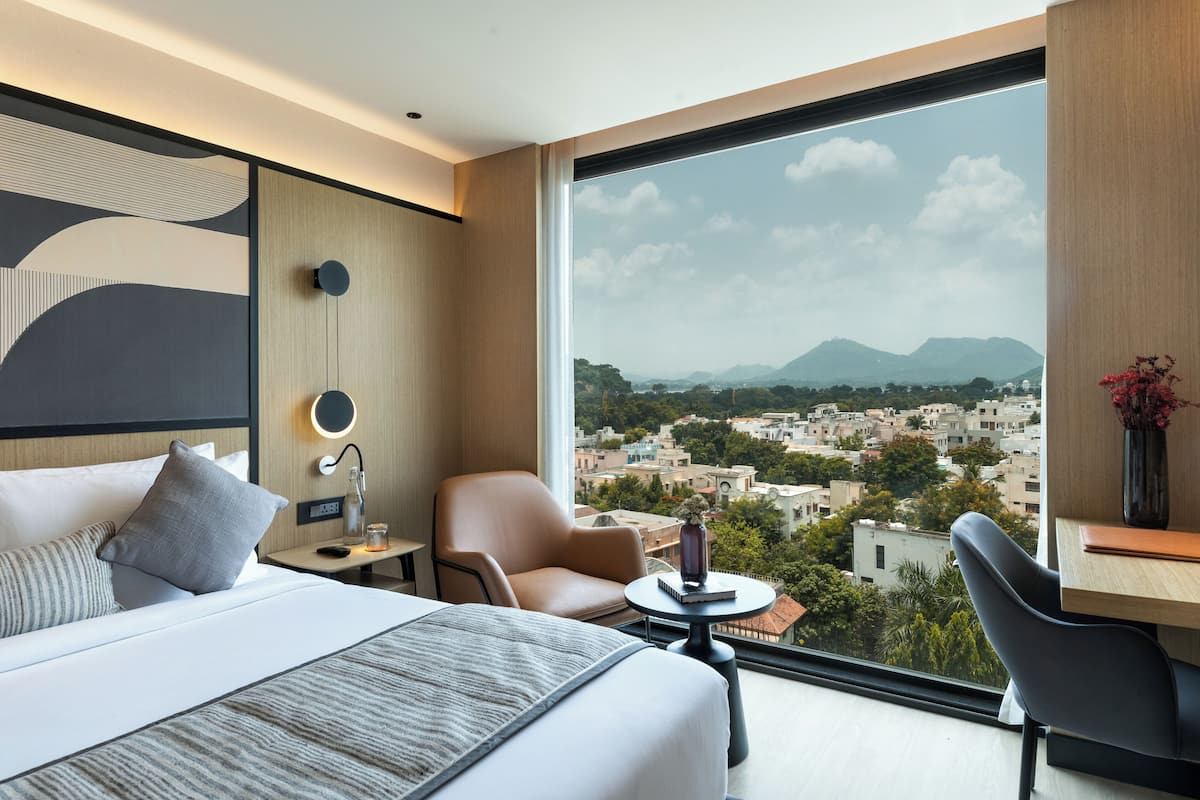 A modern hotel room with a large bed, a desk, a chair, and a view of a cityscape through a large window.