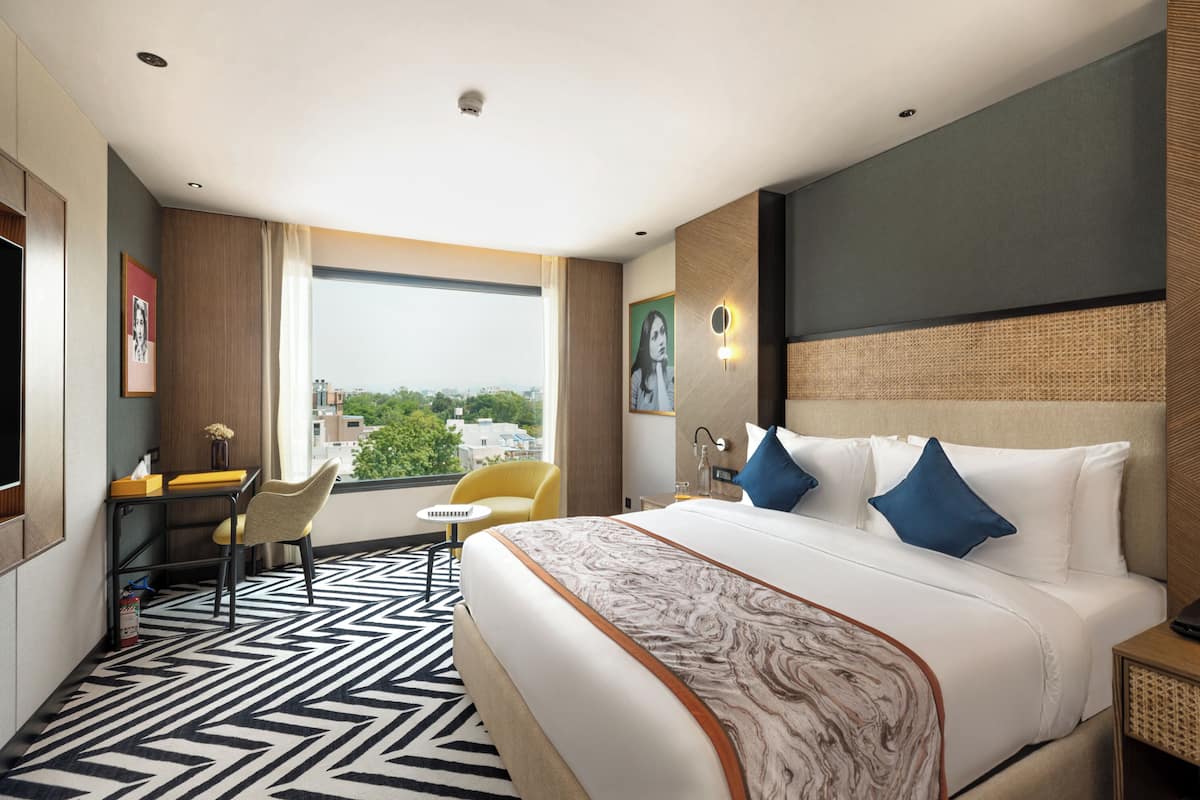 A hotel room with a large bed, a desk with a chair, a small table, and a view of the city through a large window.